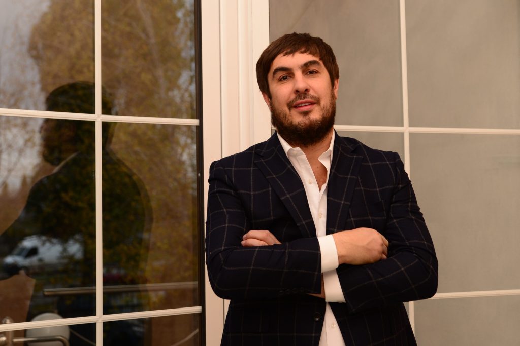 Микола Удянський з бородою в чорному піджаку та білій сорочці зі схрещеними на грудях руками
